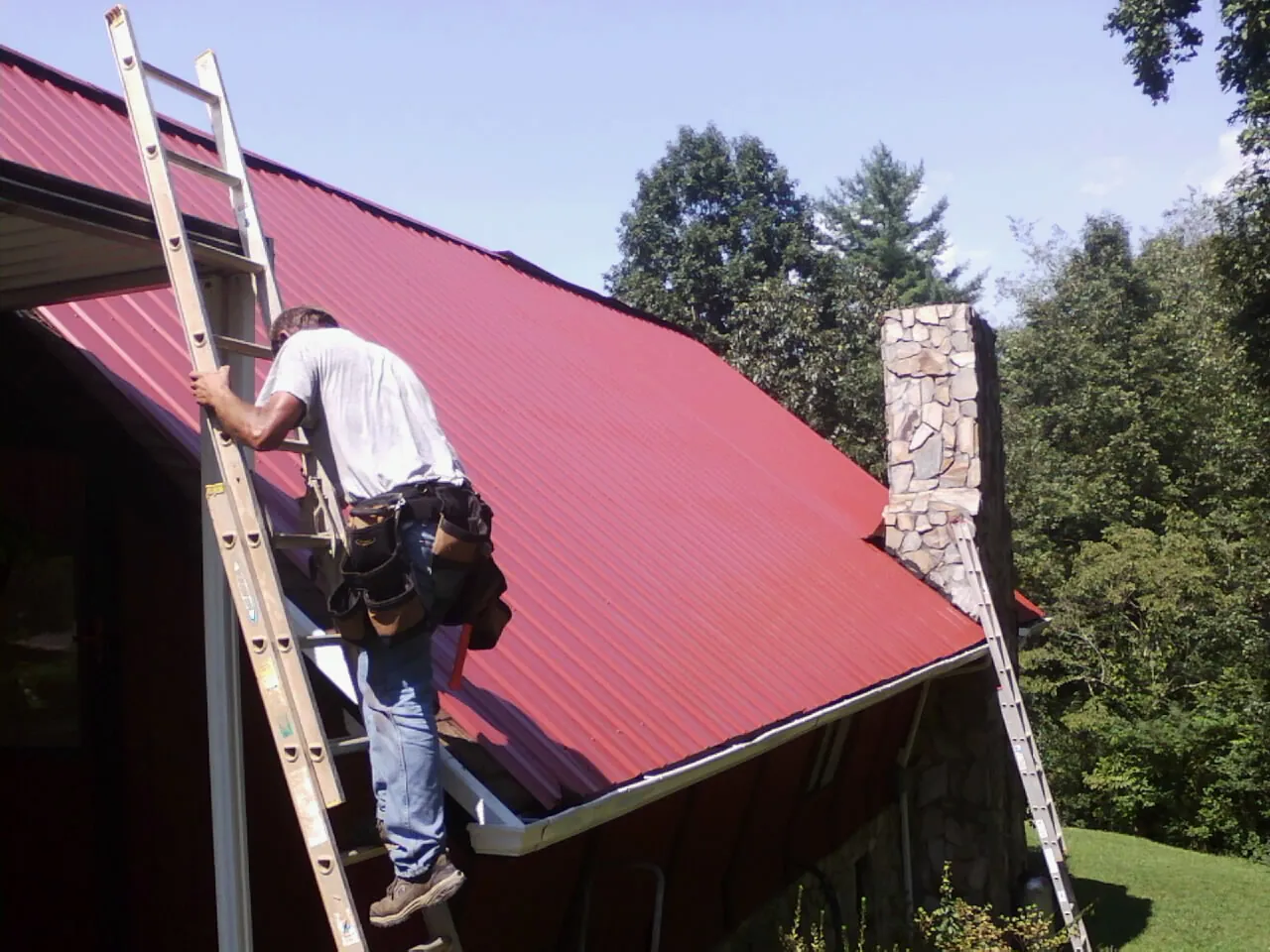 Roofer on a ladder beside a red metal roof and stone chimney — Soffit & Fascia Repair by Roofing Brothers in Newton Center